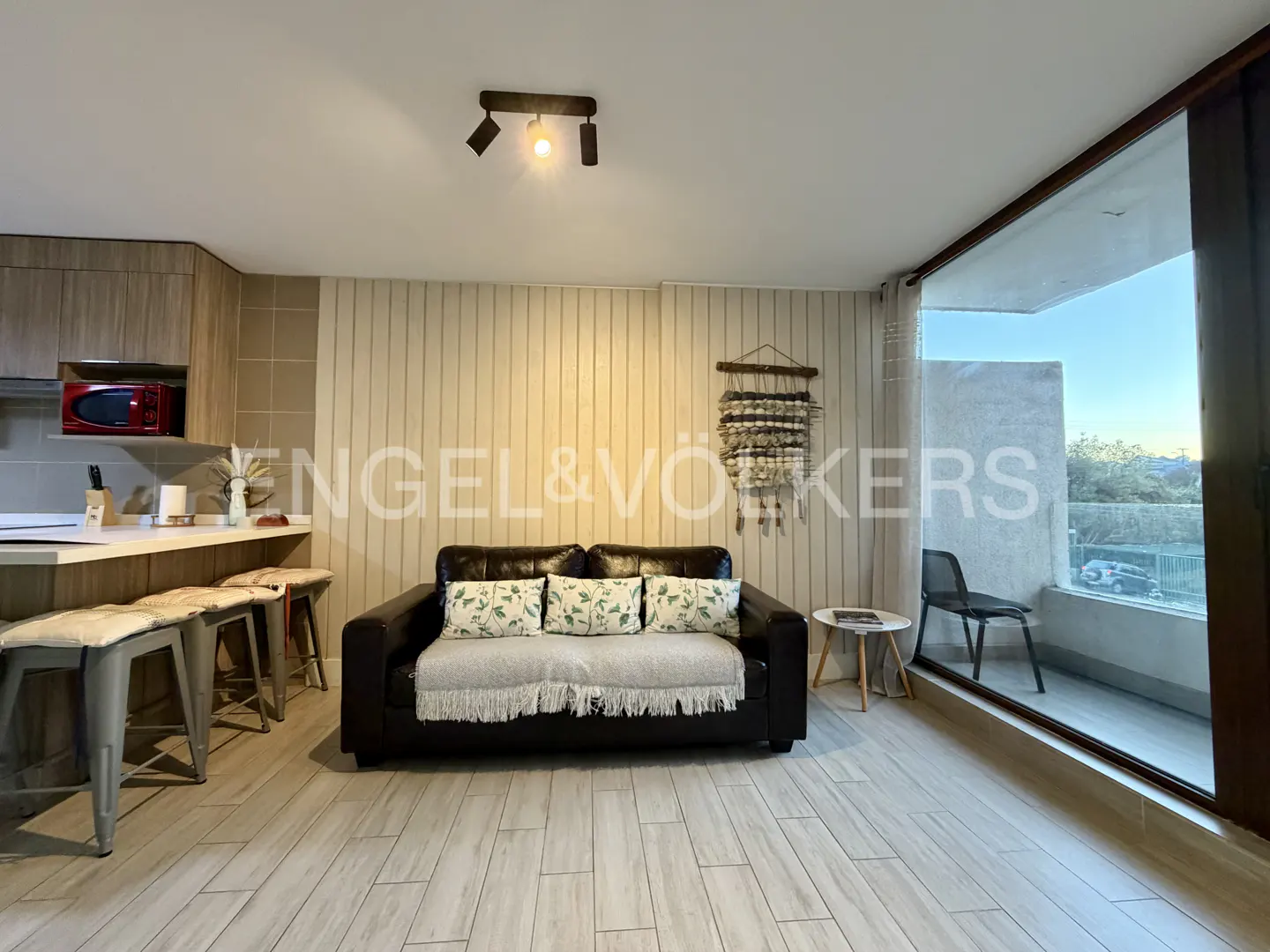Living room with a black sofa, floral pillows, and a woven wall hanging. Balcony with a chair and a view. Kitchen area with stools.