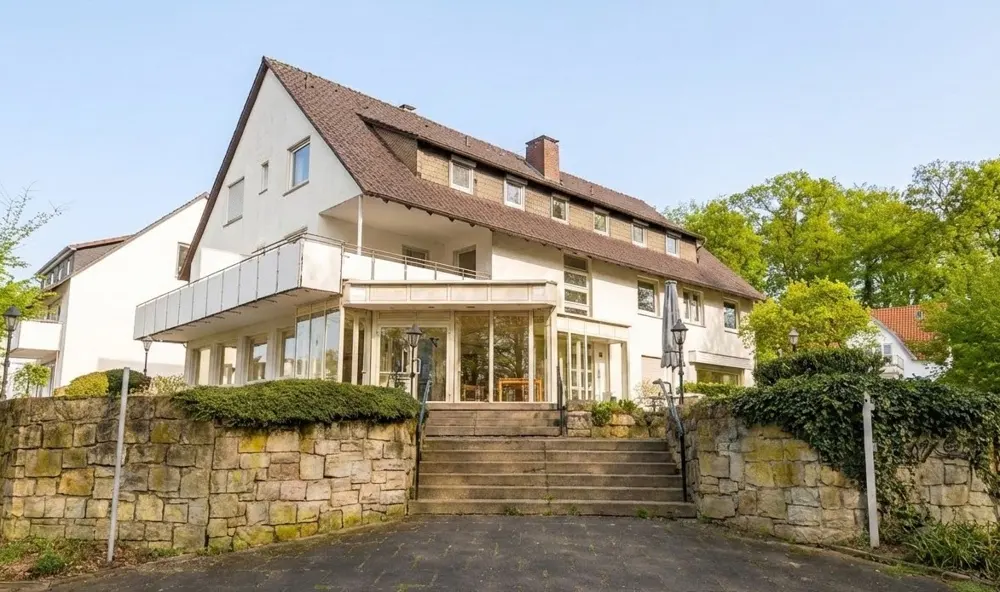 Exterior view of a white, multi-story building with a brown roof, stone walls, and a staircase leading to a glass-enclosed entrance.