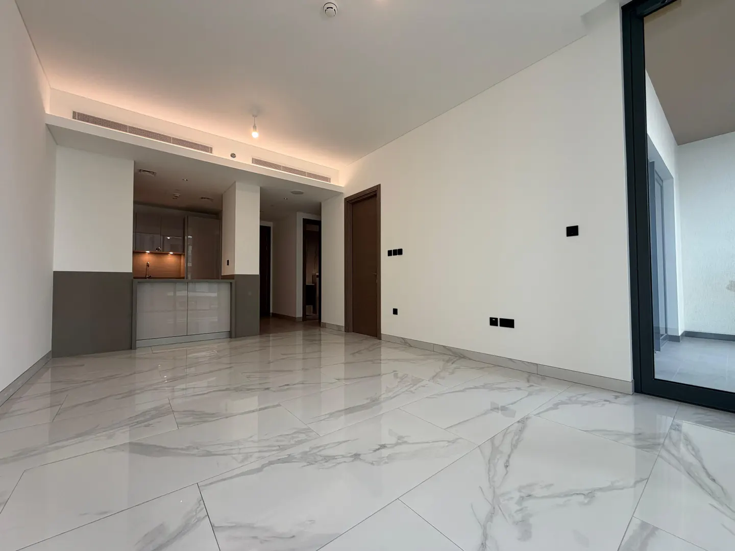 Modern living room with white marble floors, white walls, a small kitchen area, and a glass door leading to a balcony.