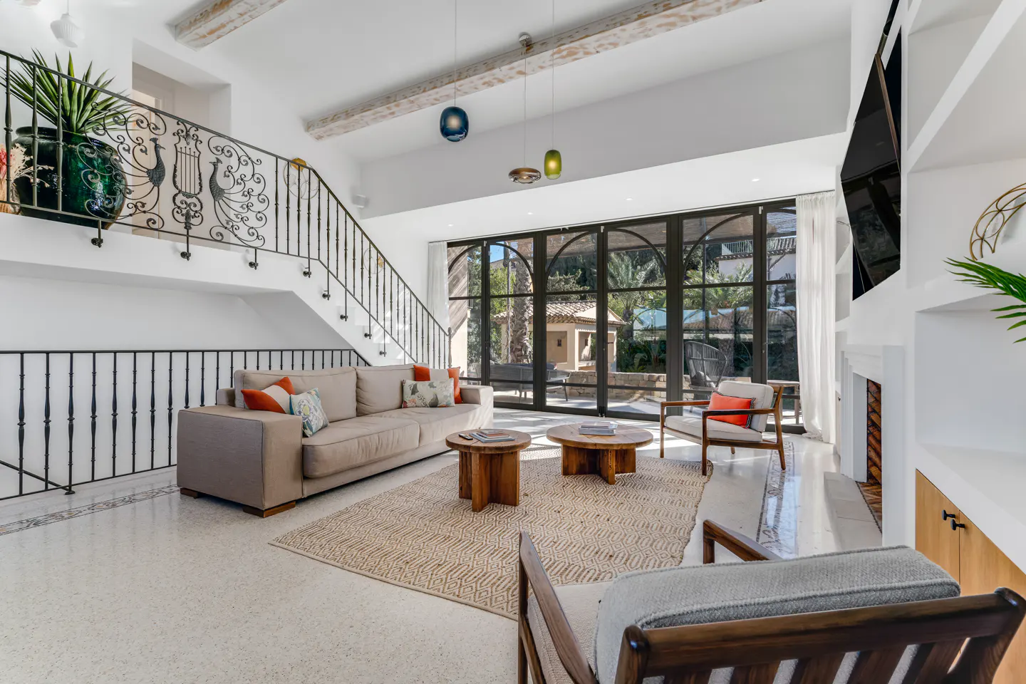 Bright living room with beige sofa, wooden tables, and chairs on a patterned rug. Black framed windows overlook a patio. Staircase with peacock railing.