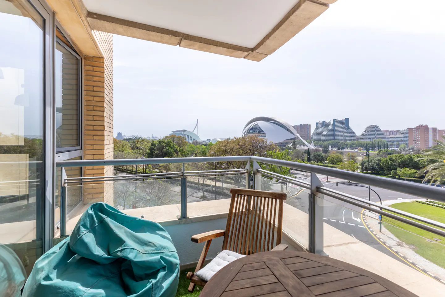 Balcony view with a teal beanbag, wooden chair, and table. Cityscape with modern architecture in the background.