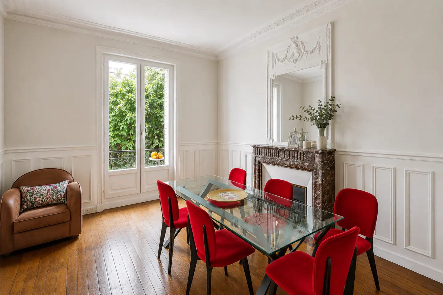 Bright dining room with glass table, red chairs, and brown armchair. Marble fireplace and white walls add elegance. View of greenery outside.