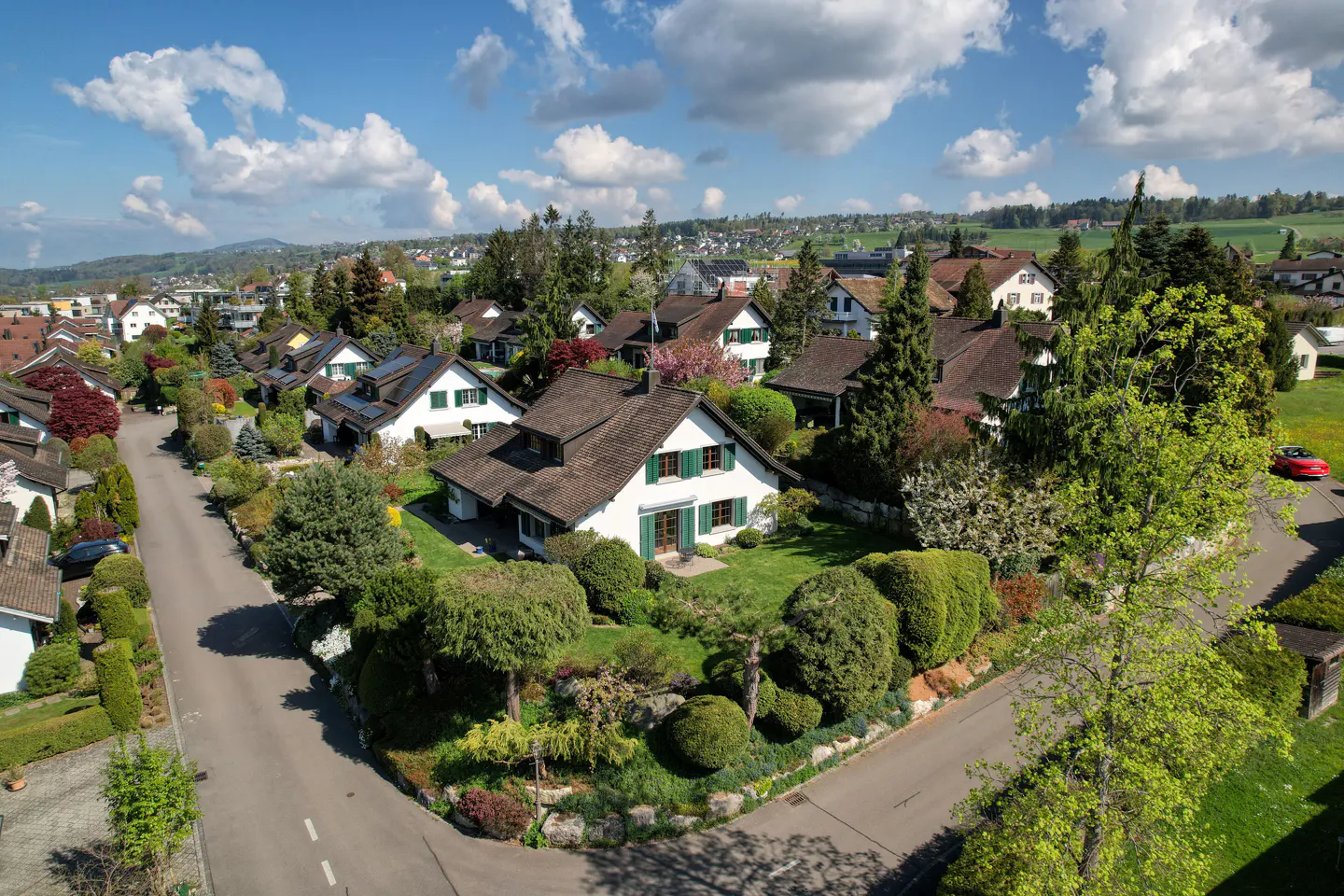 Aerial view of a suburban neighborhood with white houses, green shutters, and lush landscaping under a blue sky with clouds.