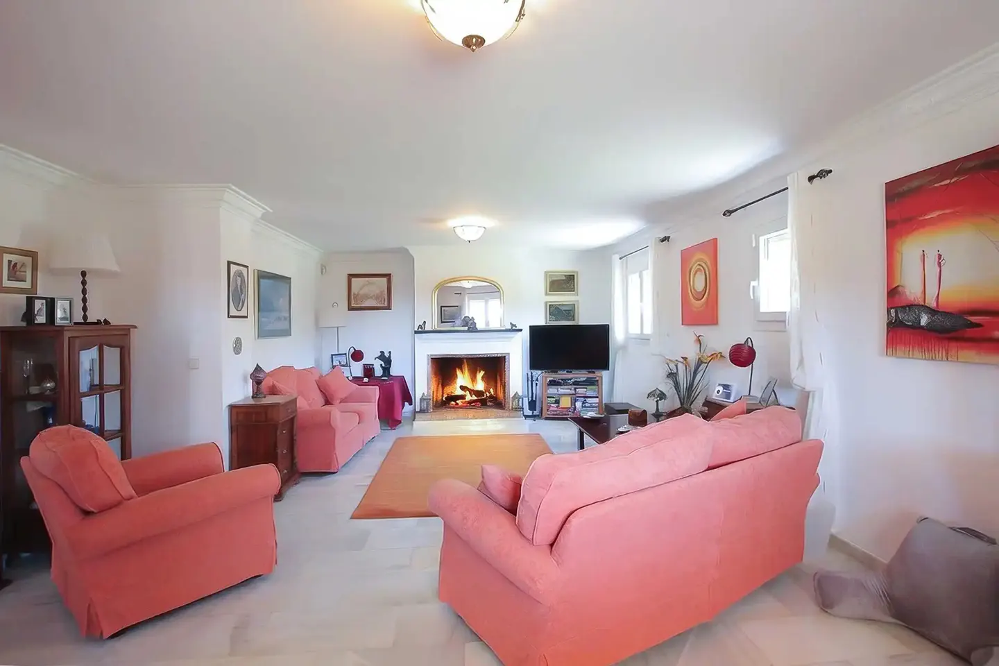 Bright living room with white walls, marble floors, and coral sofas. A fireplace with a burning fire is centered. Artwork adorns the walls.