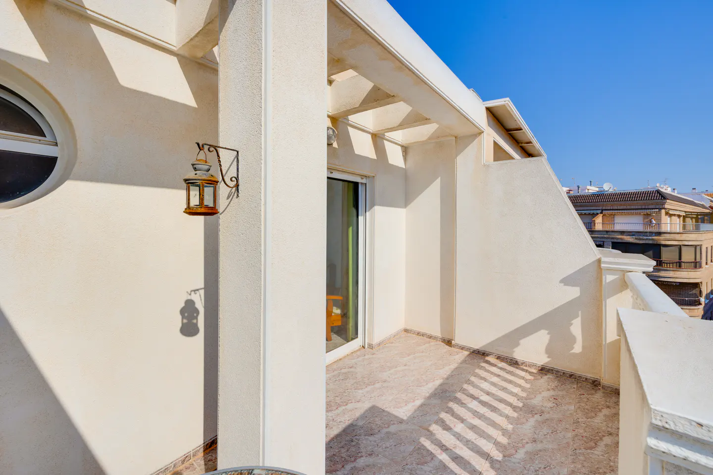 Sunny cream-colored balcony with a decorative wall lantern, tiled floor, and a view of neighboring buildings under blue sky.