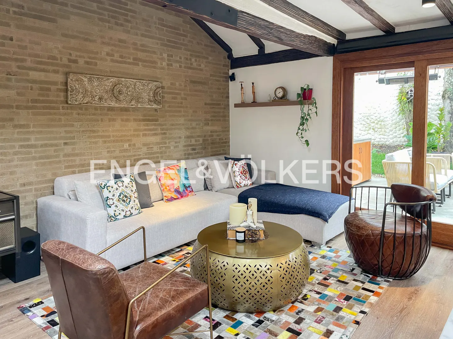 Living room with a light gray sofa, brown leather chairs, and a gold coffee table on a colorful patchwork rug. Brick wall and exposed beams.