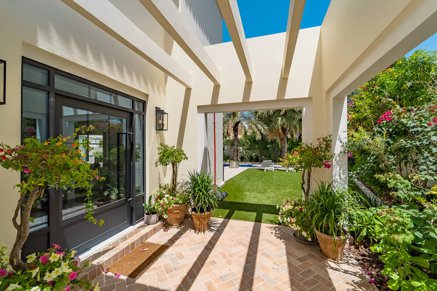Covered patio with brick floor, potted plants, and a view of a green lawn, pool, and lounge chair. Cream-colored walls and pergola.