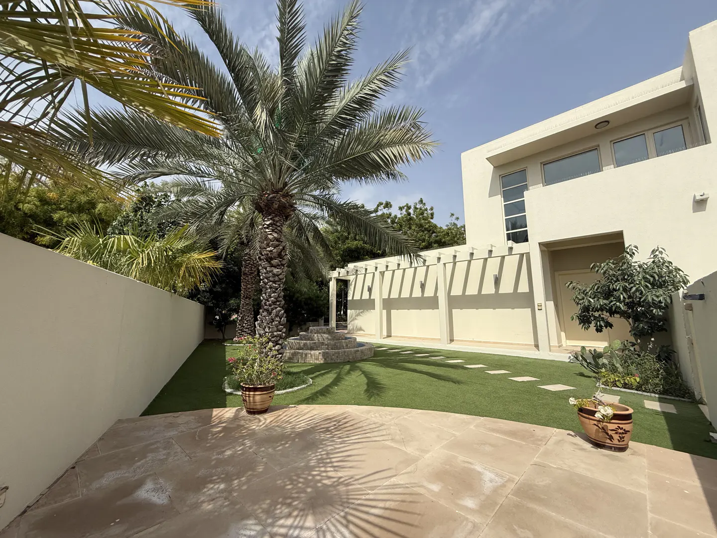 Backyard view of a modern beige house with a green lawn, palm trees, and a stone patio under a blue sky.