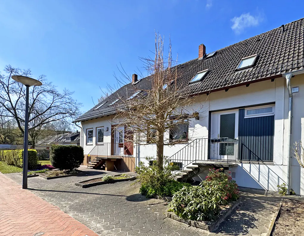 Exterior view of a two-story white townhouse with a dark gray roof and dormer windows, set against a clear blue sky. A bare tree stands in front.