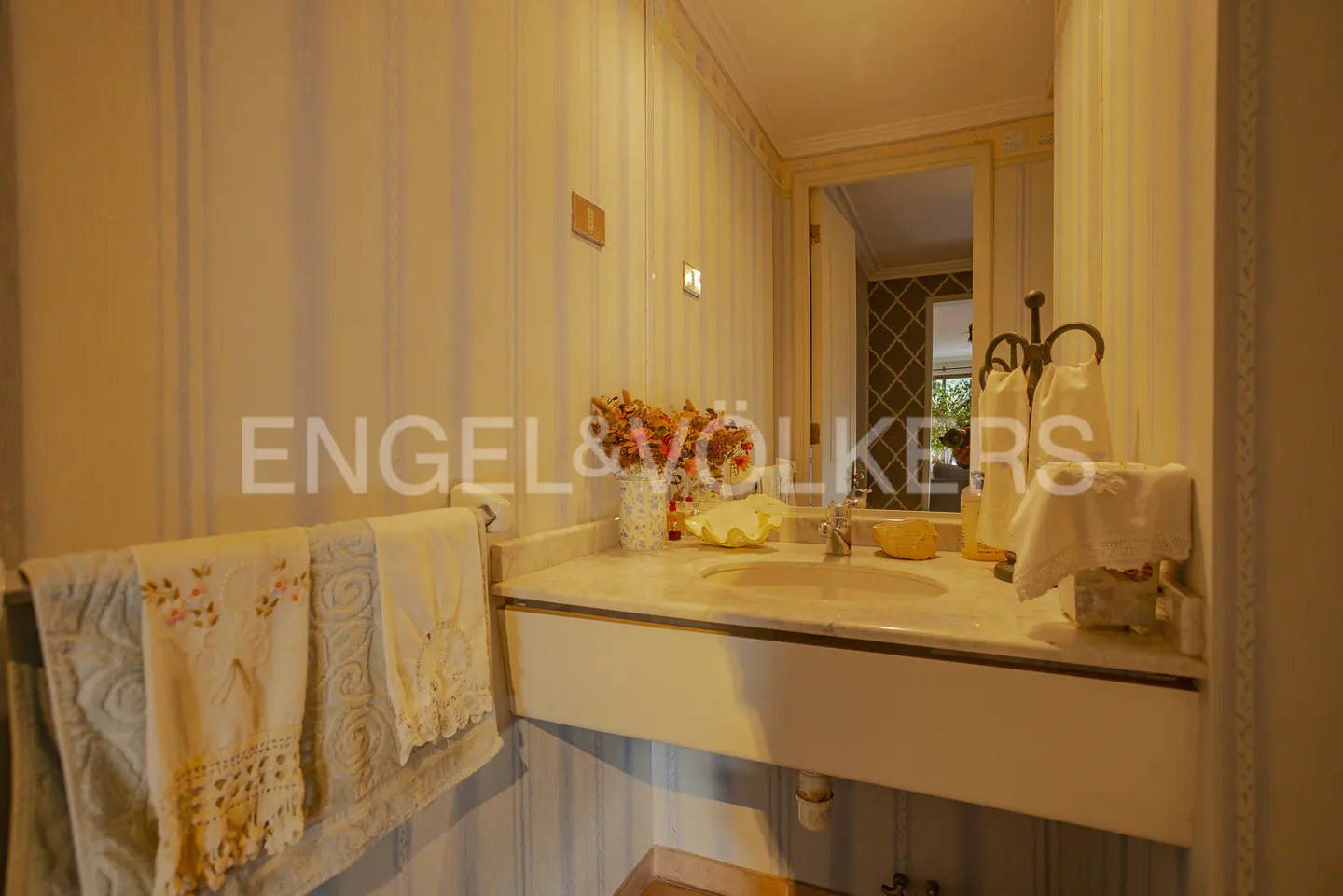 Bathroom with marble sink, mirror, and striped wallpaper. Towels hang on a rack. Flowers in a vase add a touch of color.