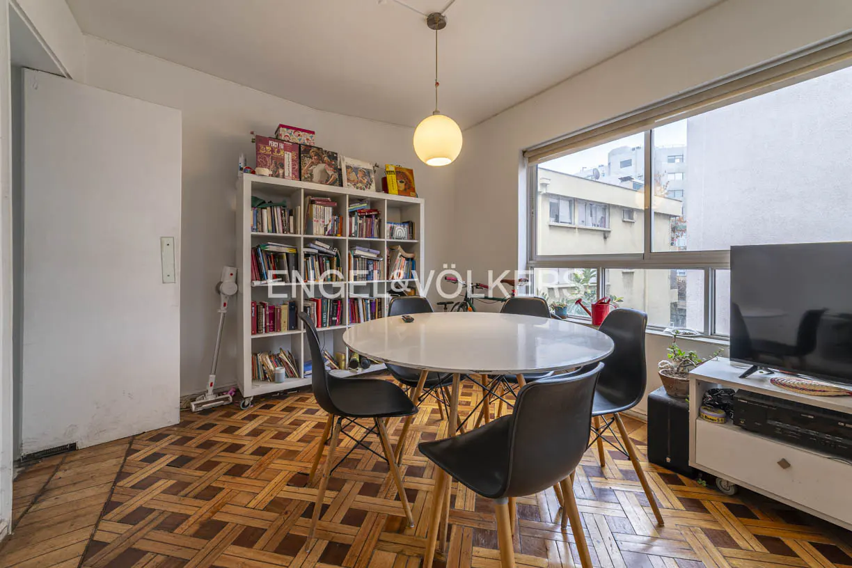 Dining area with a round white table, four black chairs, a white bookshelf, and a large window on a parquet wood floor.