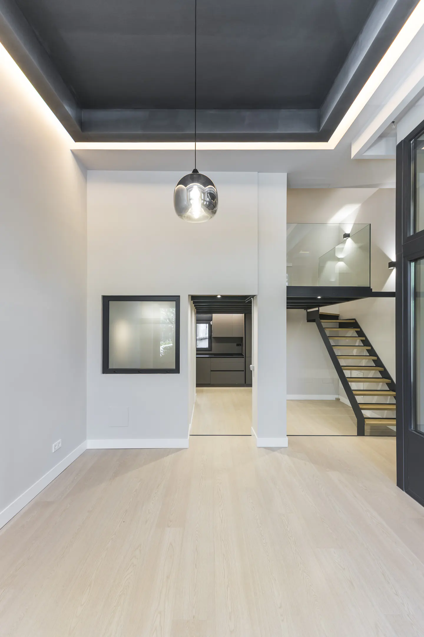 Modern open-plan living area with light wood floors, a dark tray ceiling, a glass pendant light, and a floating staircase.