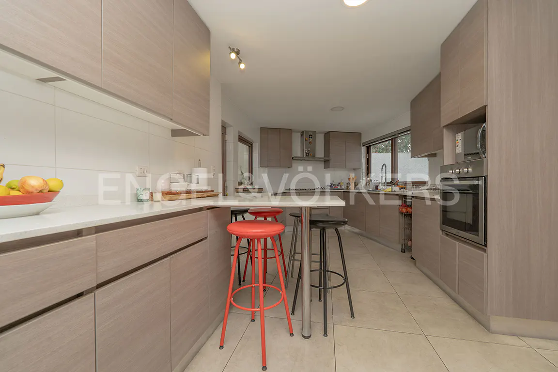 Modern kitchen with light wood cabinets, white countertops, a breakfast bar with red and black stools, and built-in oven.