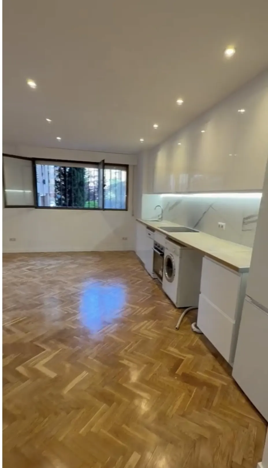 Modern kitchen with white cabinets, built-in appliances, and light wood herringbone floors in an open-plan living space.