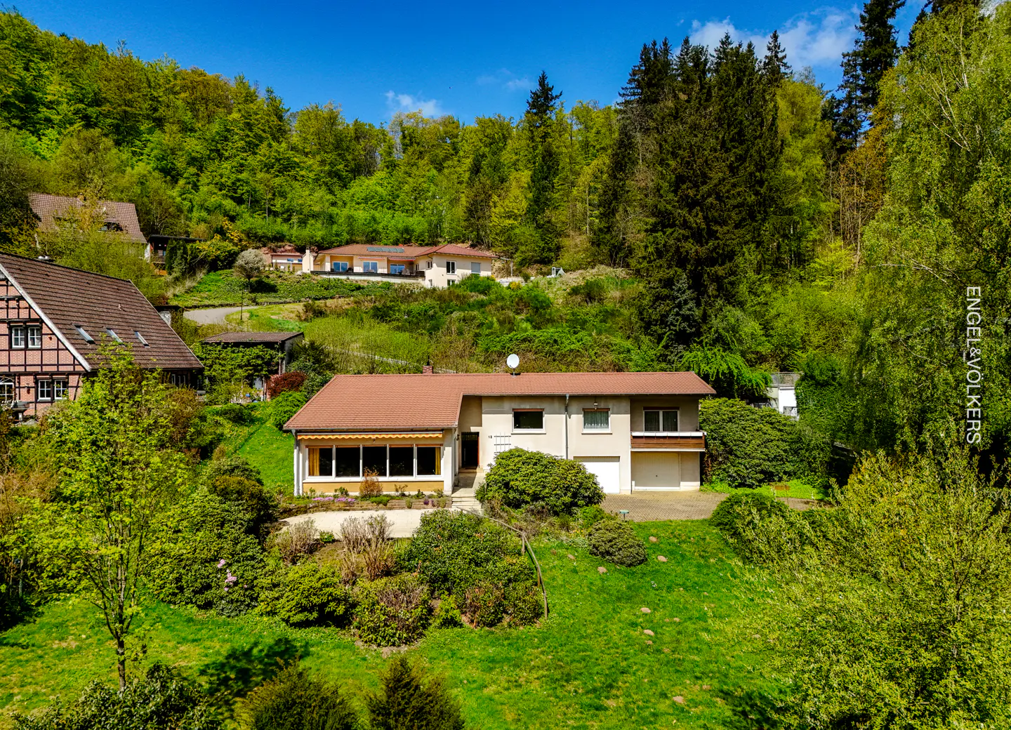 A beige house with a brown roof sits on a green lawn, surrounded by lush green trees and a blue sky.