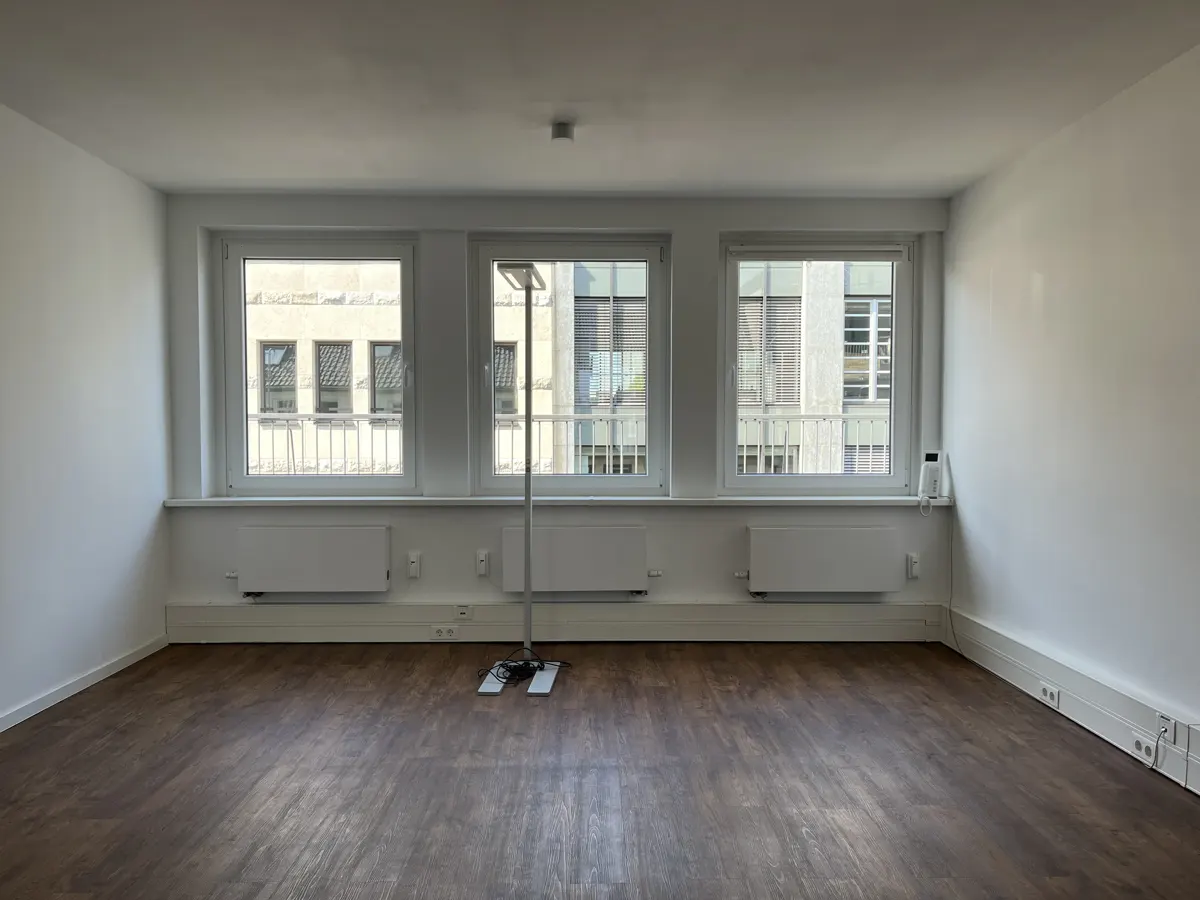 Empty white room with three windows, a brown floor, and white walls. Radiators are under the windows. A light stand is in the center.