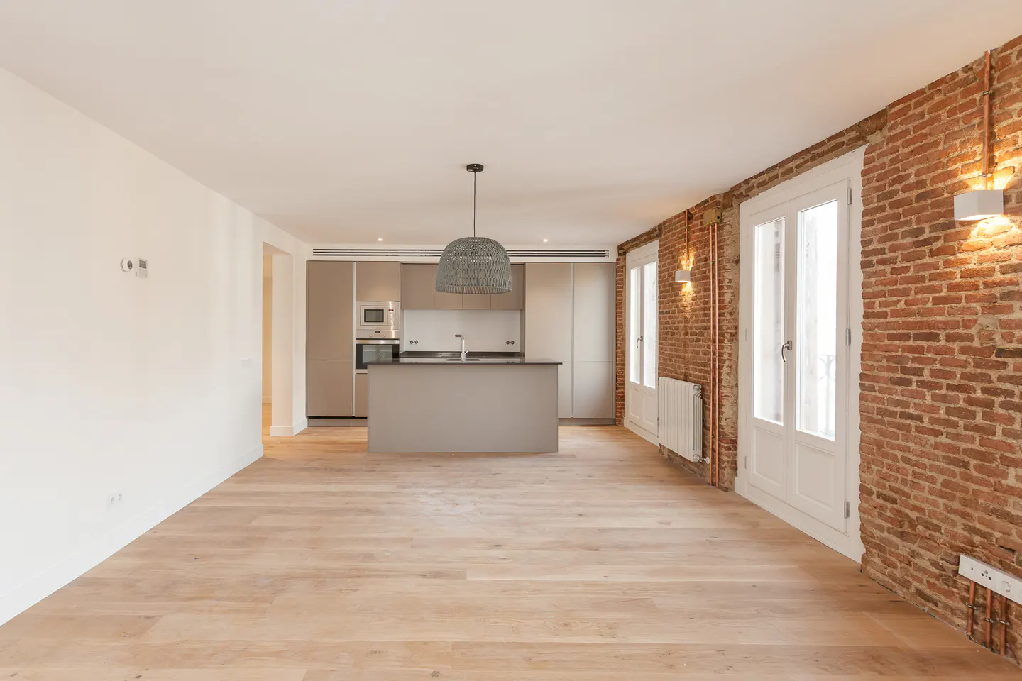Bright, open apartment with hardwood floors, exposed brick wall, and a modern kitchen with island. Woven pendant light hangs above.
