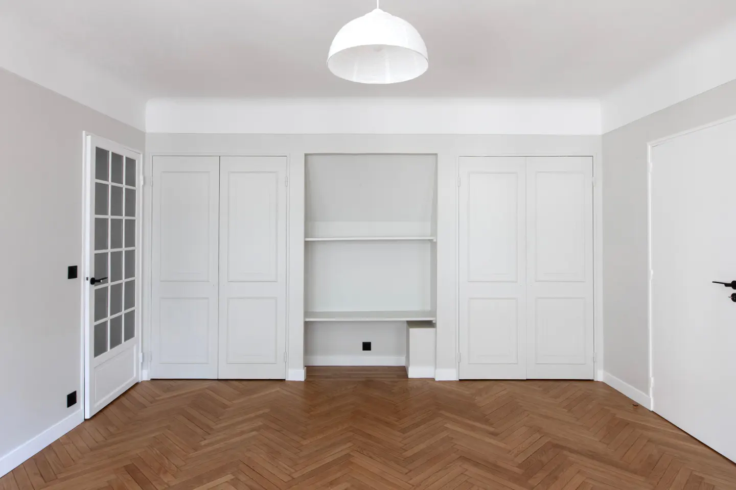Minimalist room with herringbone wood floors, white walls, built-in closets, open shelving, and a white pendant light.