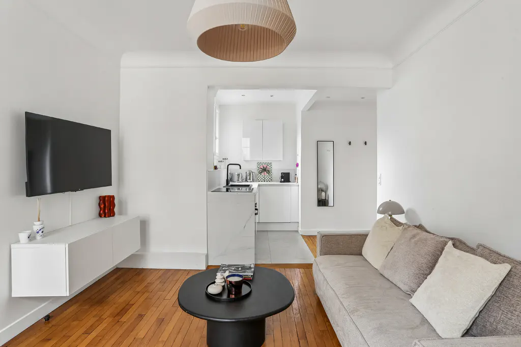 Modern living room with hardwood floors, a grey sofa, black coffee table, wall-mounted TV, and an open white kitchen.