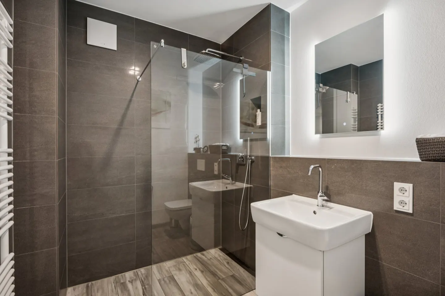 Modern bathroom with gray tile walls, wood-look floor, and a glass-enclosed shower. A white vanity and illuminated mirror add brightness.