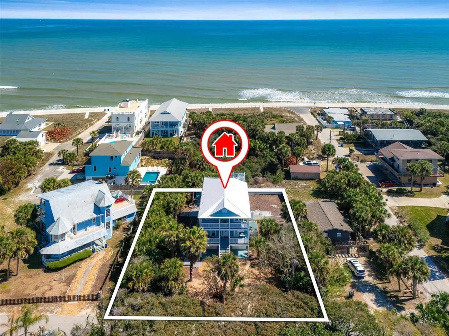 Aerial view of a blue, two-story beach house with a white roof, outlined in white, near the ocean. A red house icon marks the location.