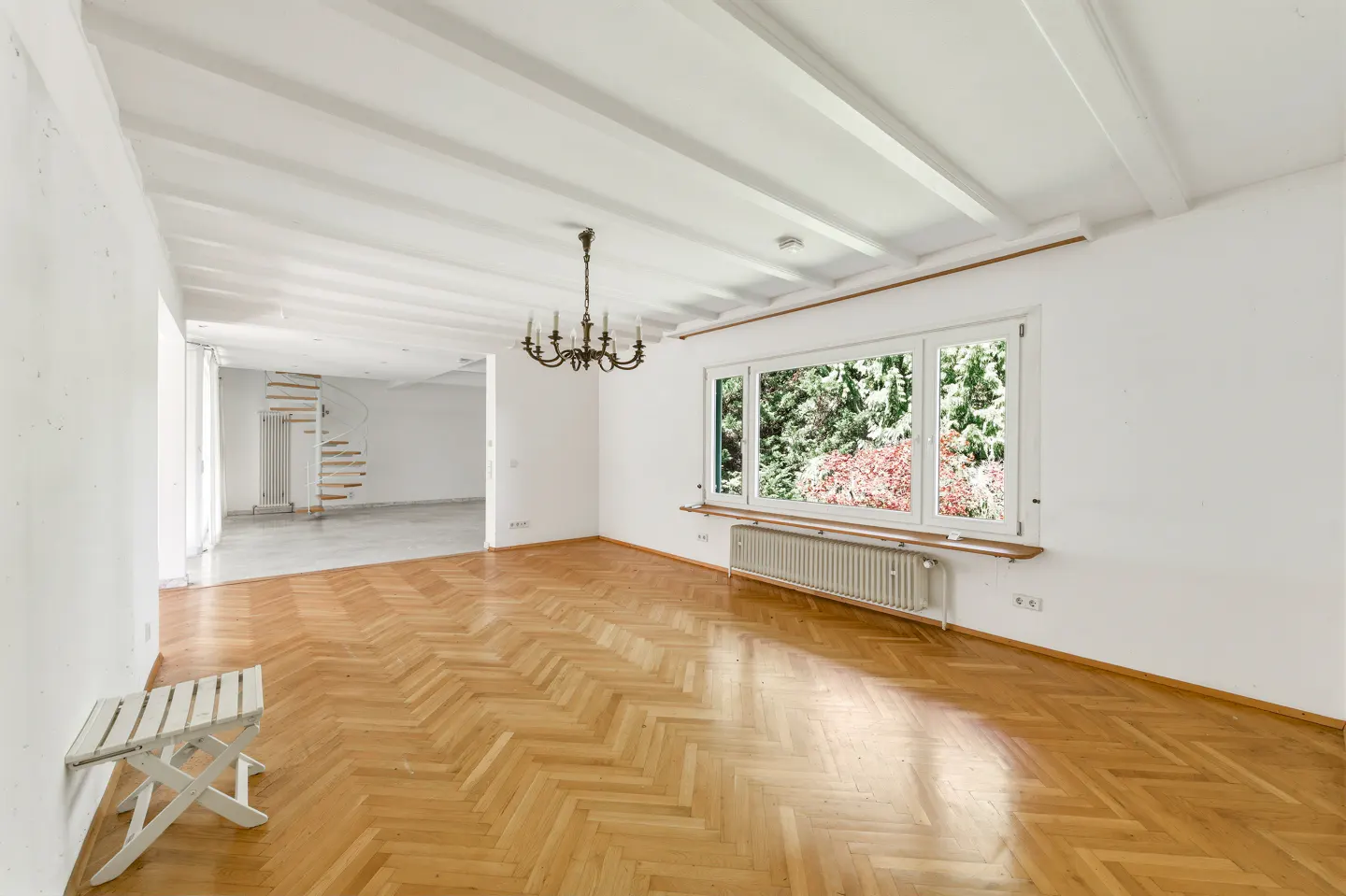 Bright, empty room with herringbone wood floors, white walls, and a large window overlooking trees. A chandelier hangs from the beamed ceiling.