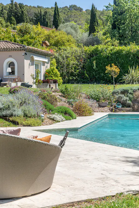 A backyard with a pool, a beige house, and lush greenery. A wicker chair sits on the stone patio.
