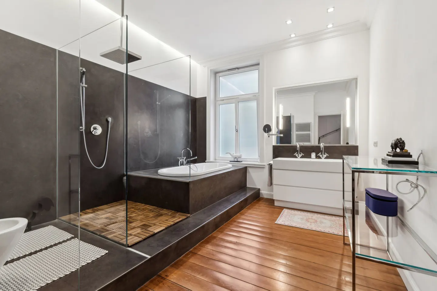A modern bathroom with a glass-enclosed shower, a white tub on a raised platform, and a white vanity with a large mirror. The floor is wood.
