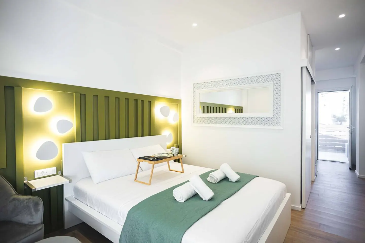 Bright bedroom with white walls, a green headboard with lights, and a white bed with a green runner and towels. A tray with items sits on the bed.