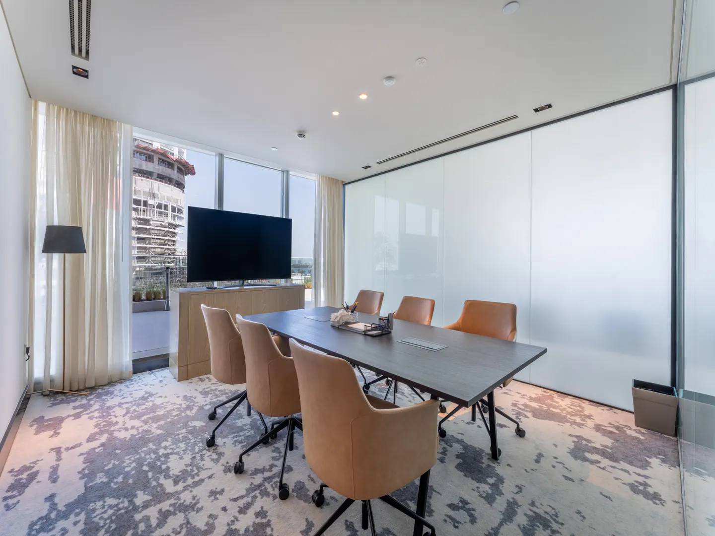 Bright conference room with a dark table, tan chairs, and a large TV. A building is visible through the window.