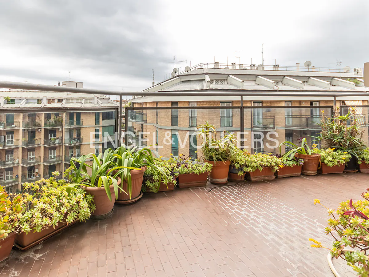 A rooftop terrace with potted plants, a brick floor, and a city view under a cloudy sky.