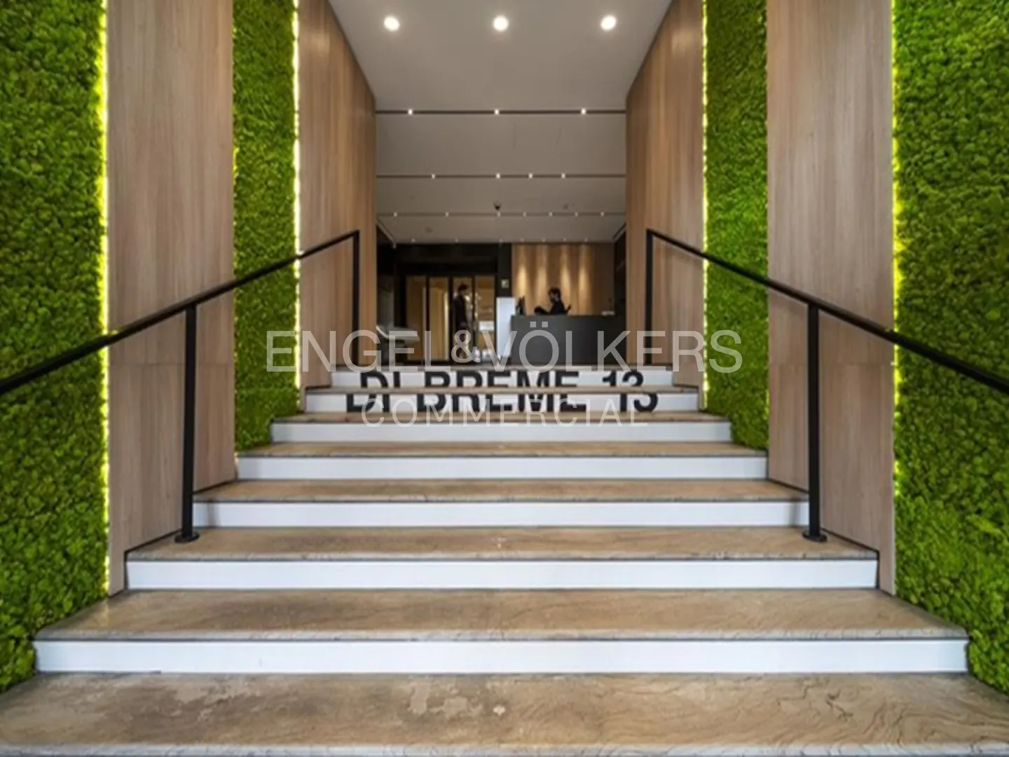 Entrance to Engel & Volkers office with stairs, black railings, and green moss walls. Reception desk visible inside.