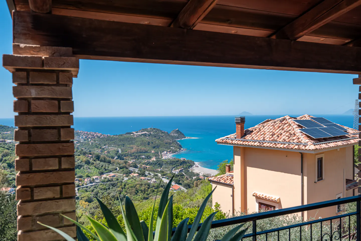 View from a covered porch of a tan house with solar panels, overlooking a green hillside, a blue ocean, and a clear blue sky.