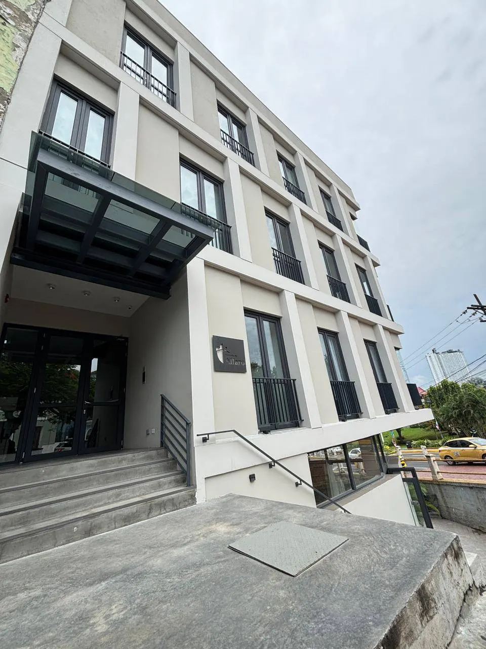 Exterior of a modern, multi-story building with a glass awning over the entrance. The building is light gray with black window frames and balconies.