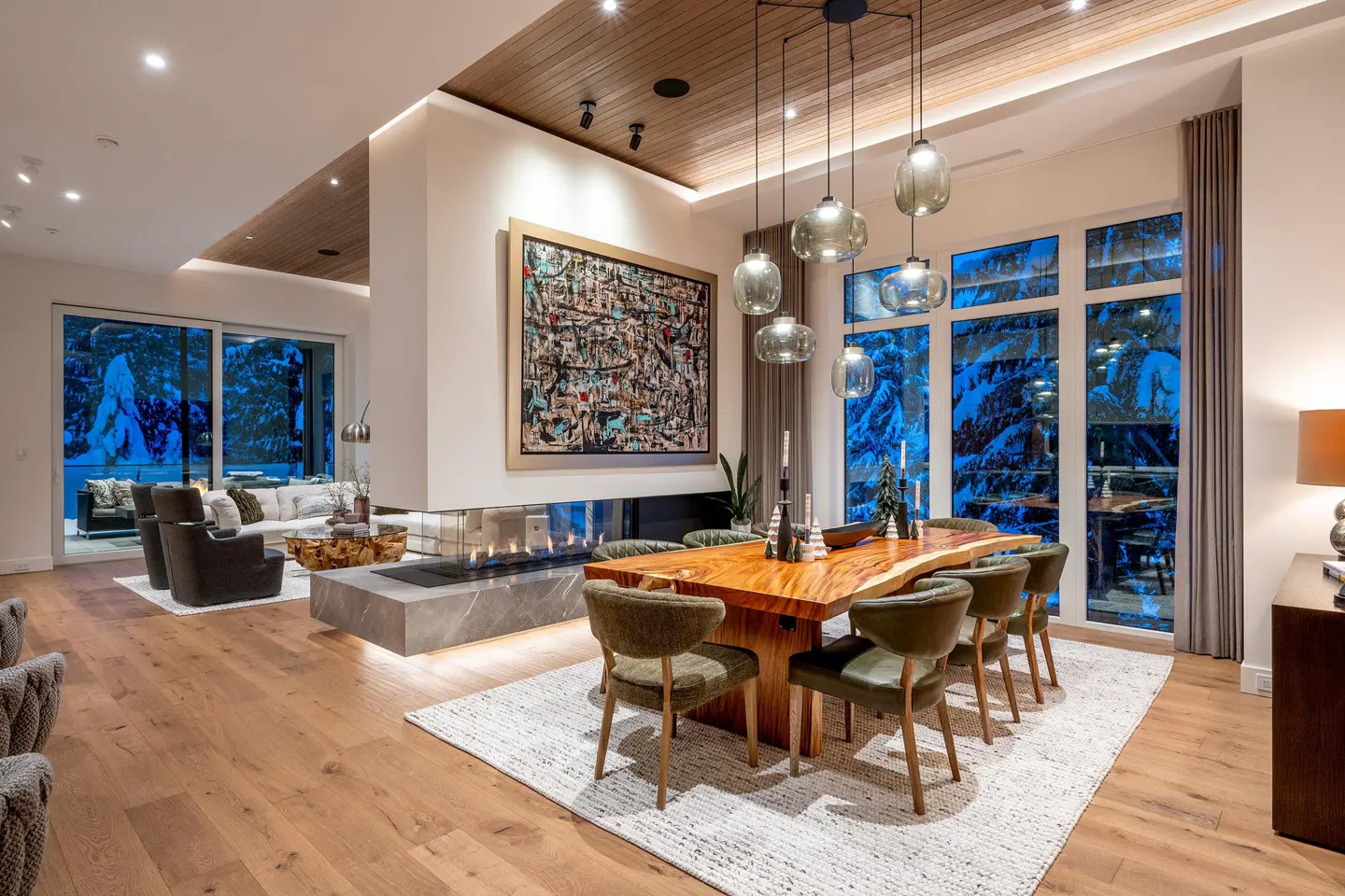 Open-concept living and dining area with wood floors, a fireplace, and a large wood table with green chairs. Snowy mountain view.