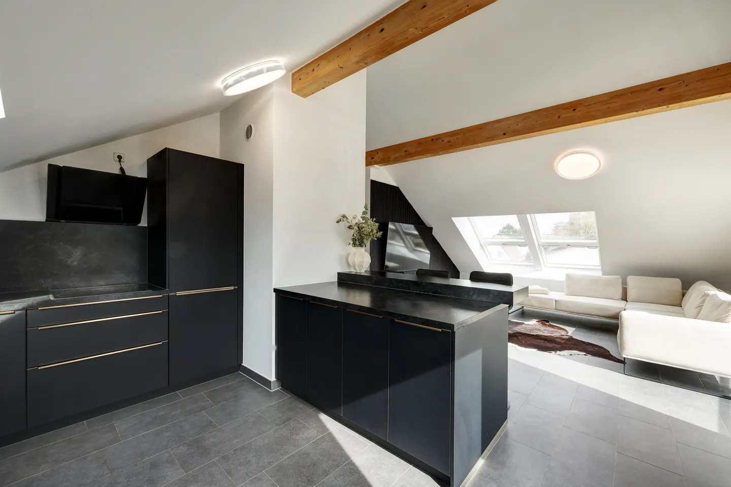 Modern kitchen and living room with black cabinets, wood beams, and white sofa in an attic apartment.