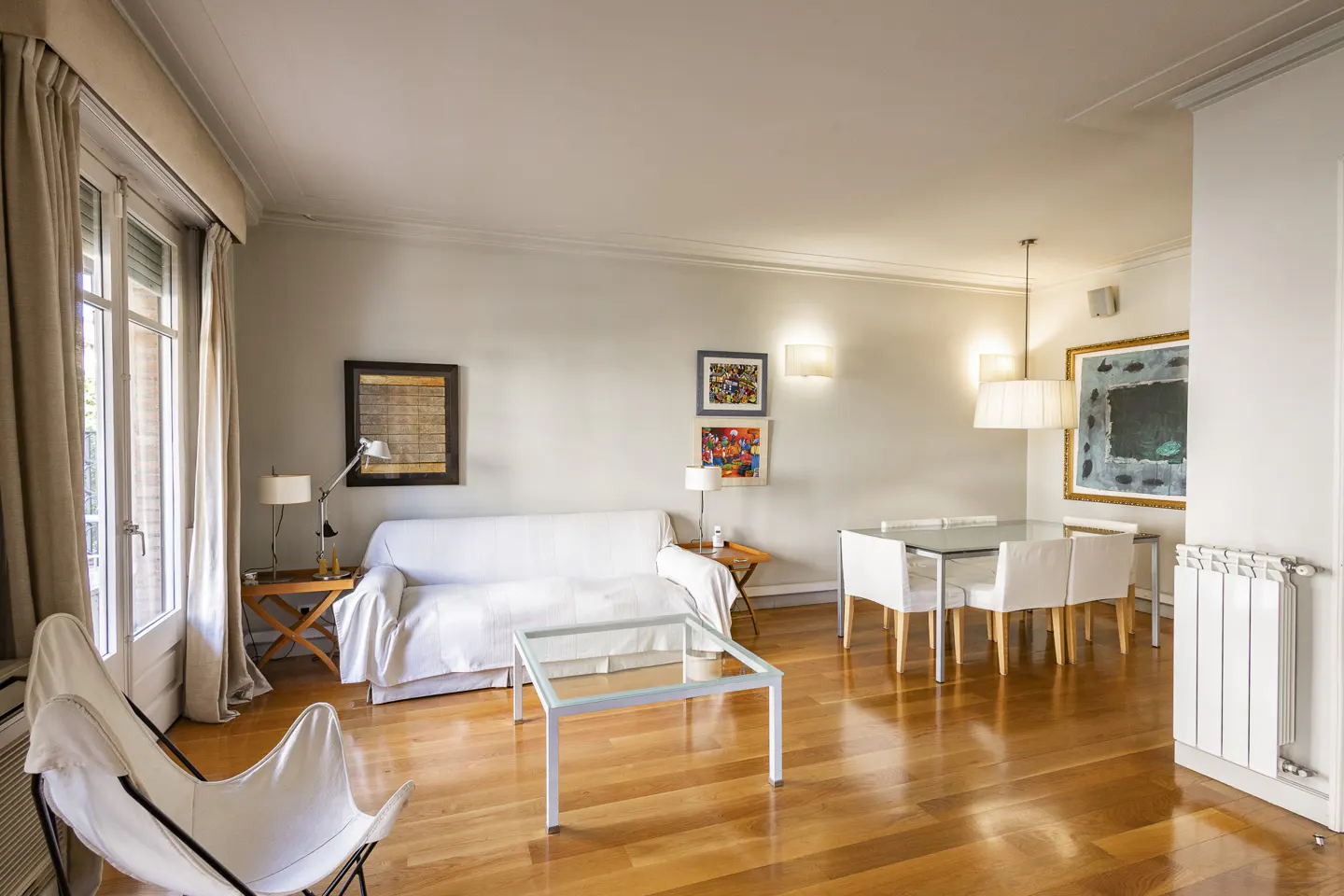 Bright living room with hardwood floors, white sofa, glass table, and dining set. Art adorns the walls. Curtains frame a window.