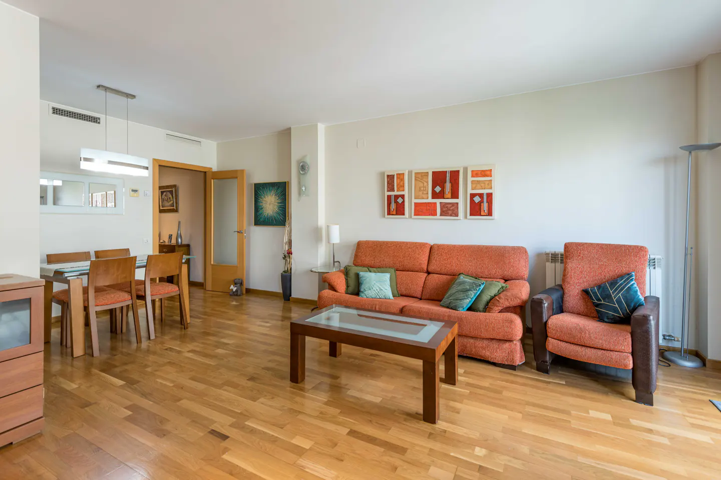Bright living room with hardwood floors, orange sofa and armchair with blue pillows, and a dining table with wooden chairs.