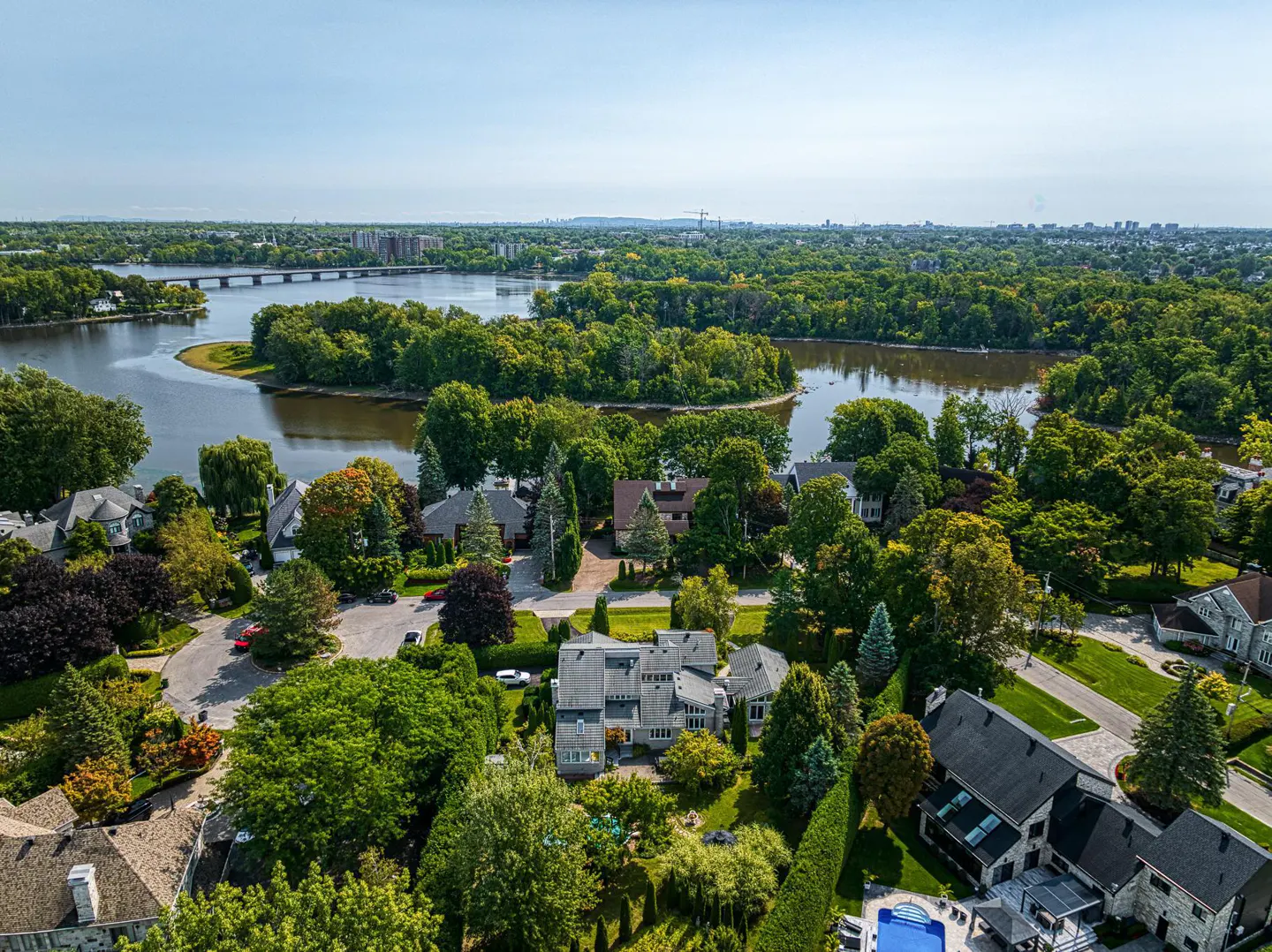 Aerial view of luxury homes nestled among lush green trees near a river and bridge on a sunny day.