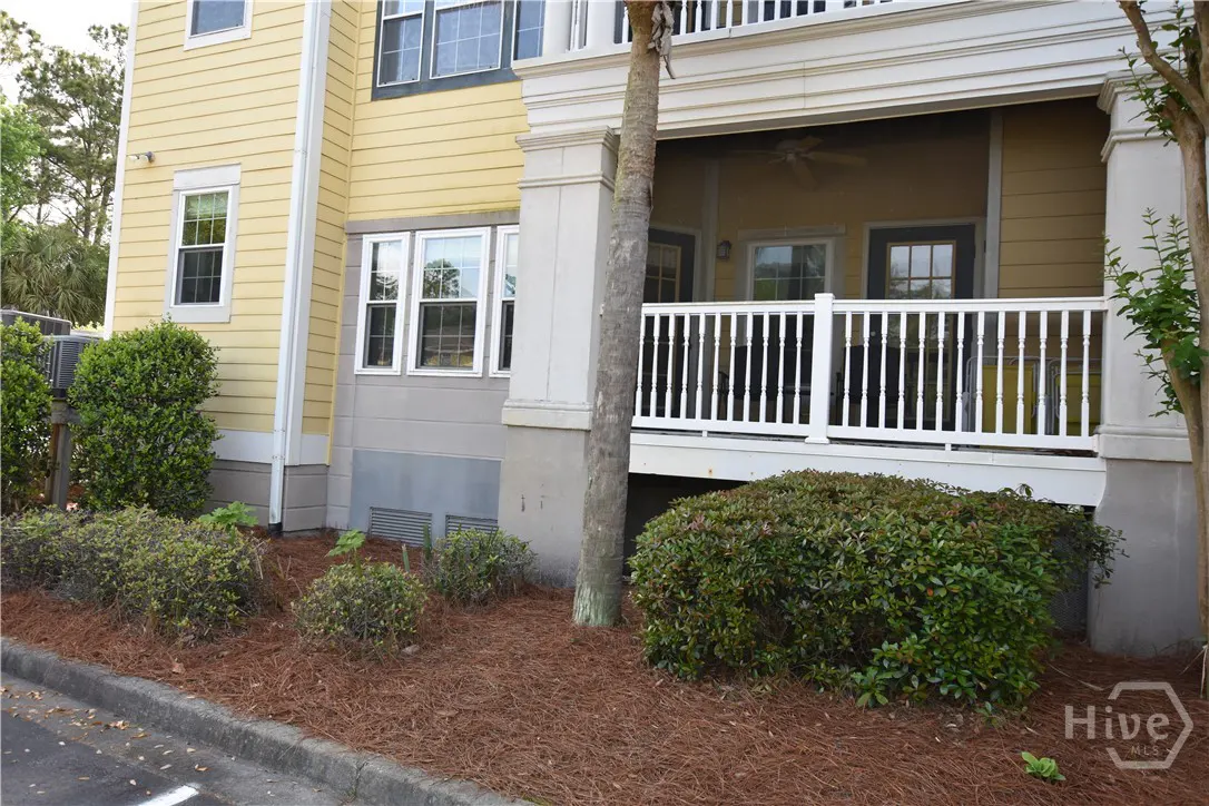 Exterior view of a yellow two-story building with a white balcony and railing. Green bushes and brown pine straw surround the base.