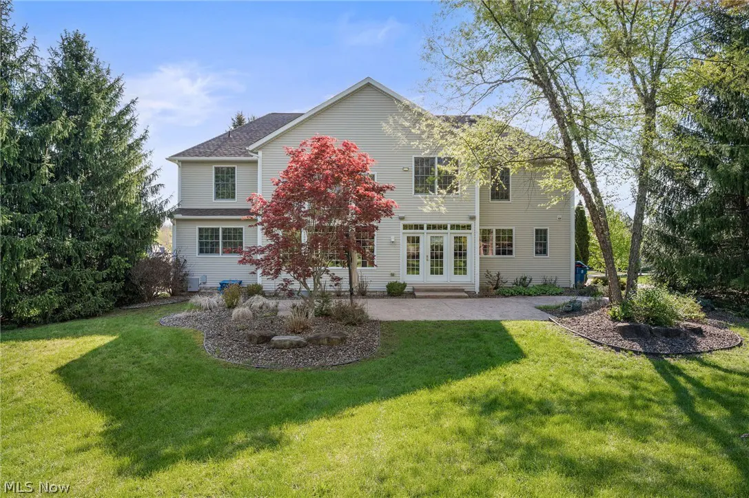 Backyard view of a two-story beige house with a patio, lawn, and trees under a blue sky. A red Japanese maple tree is centered.