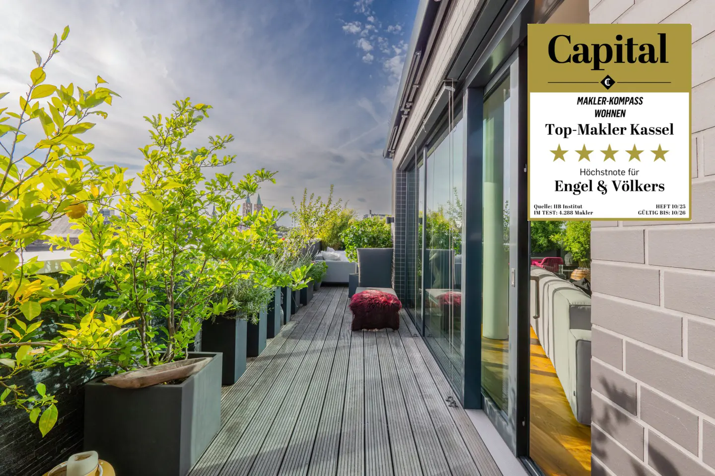 A rooftop terrace with potted plants, gray wood flooring, and a view of the city skyline under a blue sky. "Capital Top-Makler Kassel" badge on glass door.