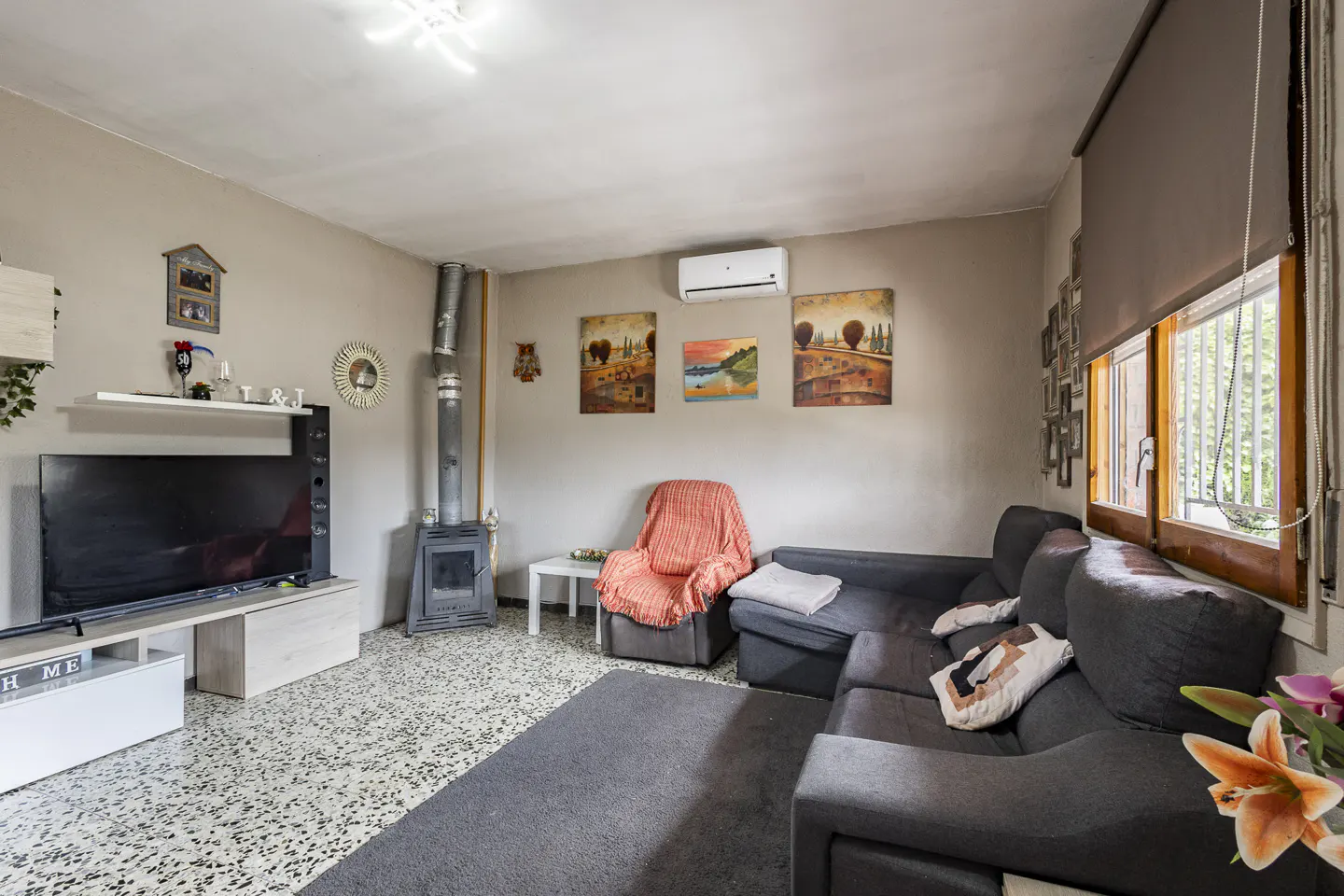 Living room with gray sectional sofa, wood-burning stove, and TV. Paintings adorn the beige walls. A window with a brown shade lets in natural light.
