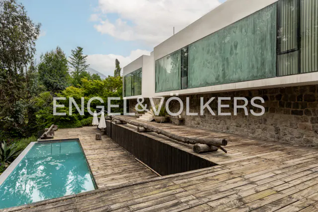 Exterior view of a modern white house with large green windows, a pool, and a wooden deck.