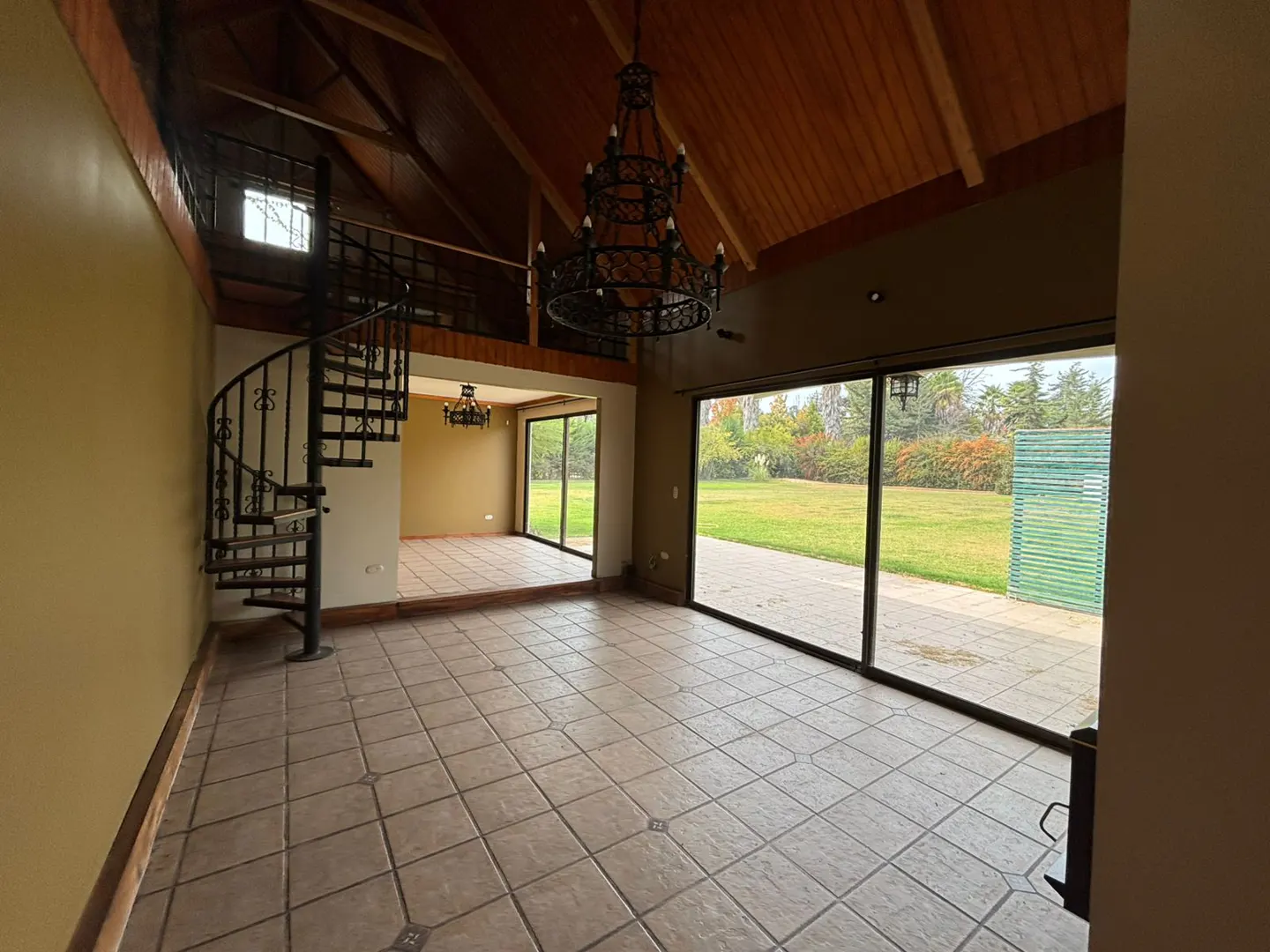 A spacious room with tile floors, a spiral staircase, and a large chandelier. Outside, a green lawn is visible through sliding glass doors.