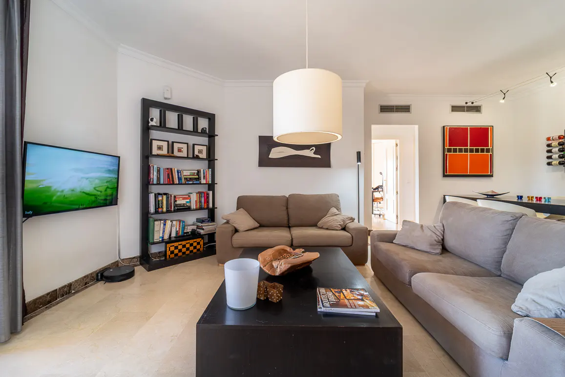 Living room with beige sofas, a dark wood coffee table, and a bookshelf filled with books. A TV hangs on the wall.