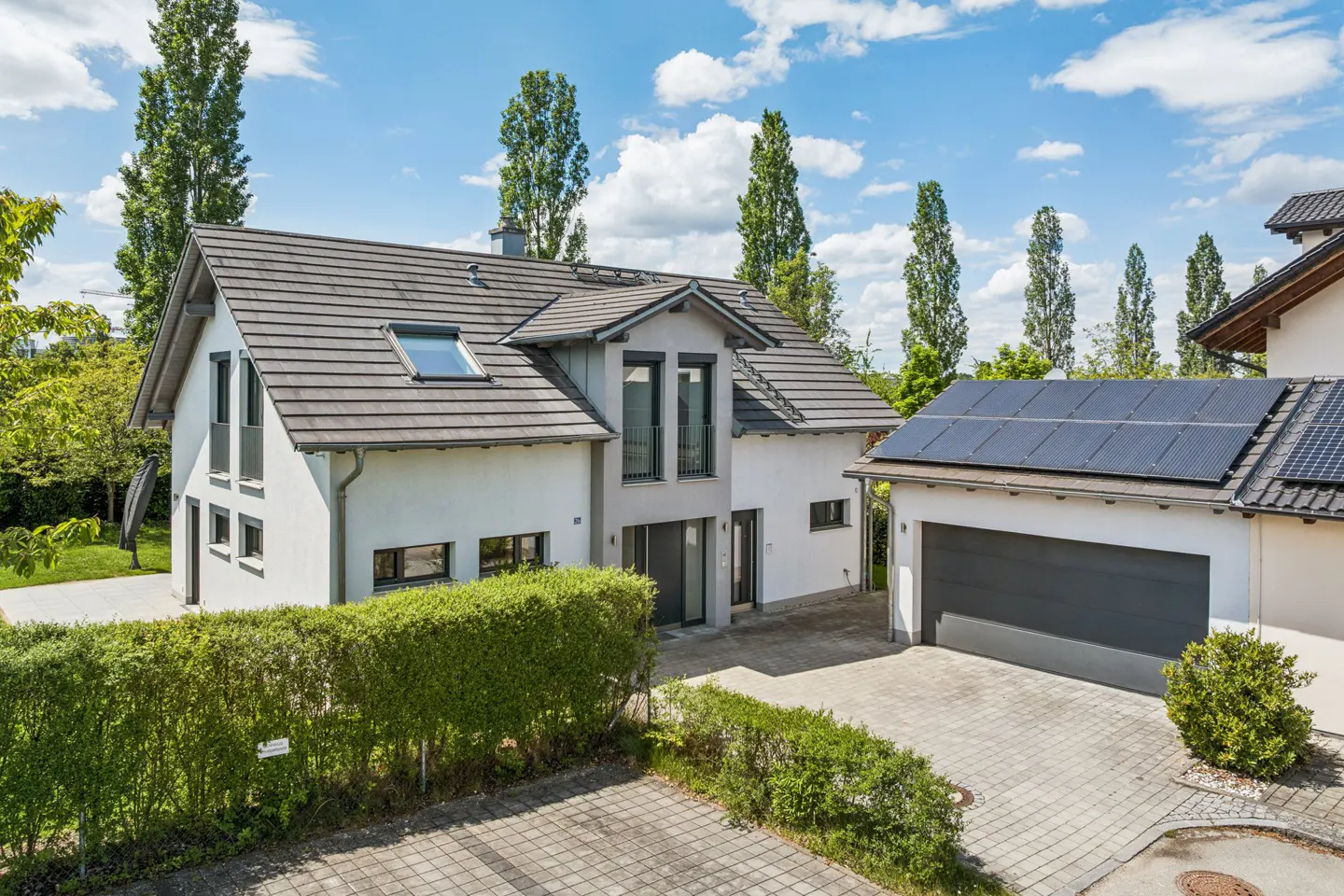 Modern white two-story house with a grey tiled roof, attached garage with solar panels, and a manicured hedge in front.