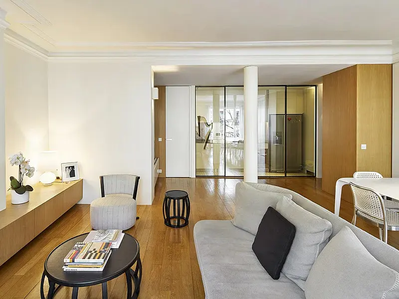 Modern living room with light wood floors, grey sofa, round coffee table, and glass-walled kitchen area in the background.