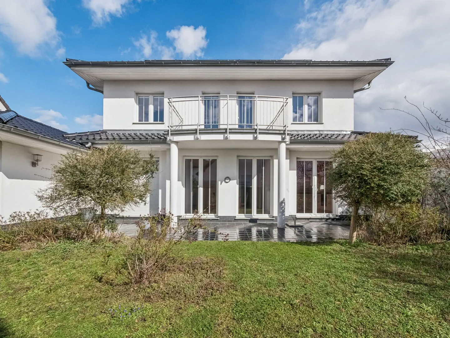 Two-story white house with a balcony, visible from the backyard with green grass and trees.