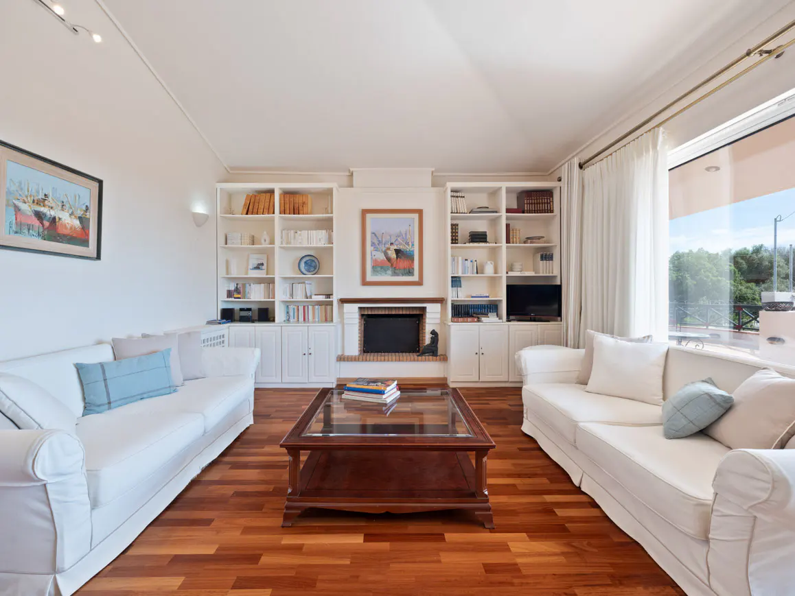 Bright living room with white sofas, wood floors, and built-in bookshelves flanking a fireplace. A glass-topped coffee table sits in the center.